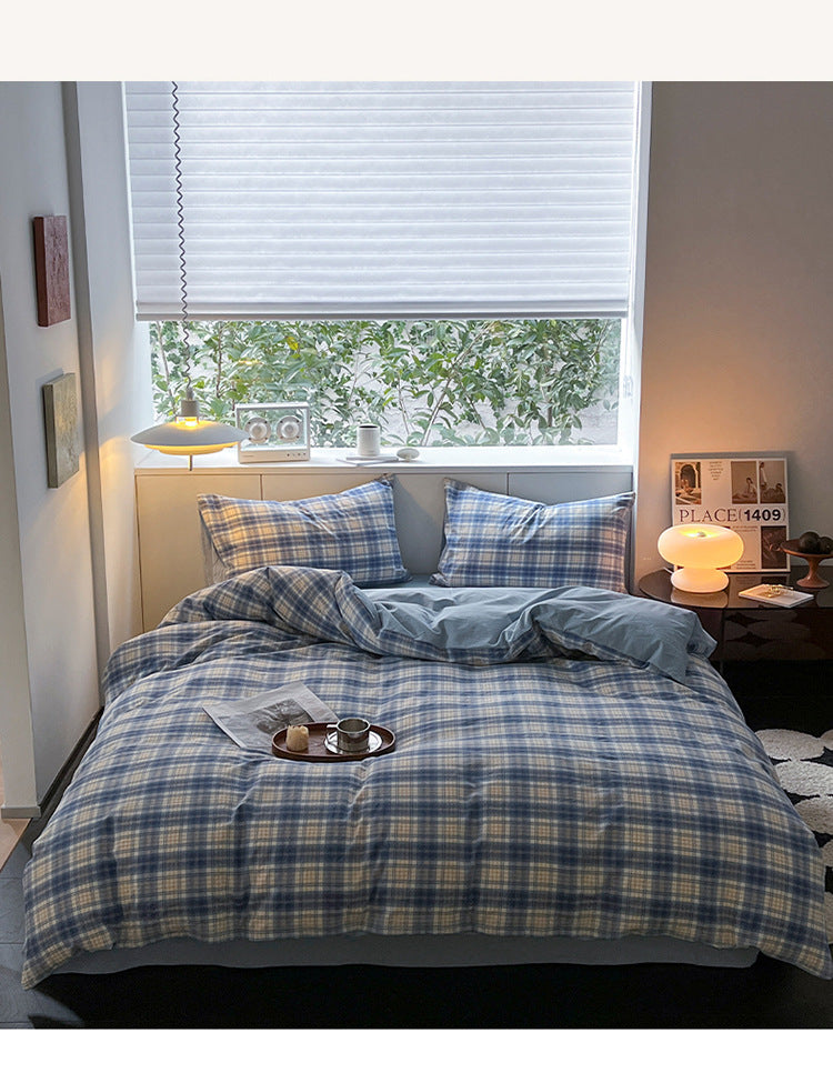 A neatly made bed in a styled bedroom setting, featuring a blue and beige plaid bedding set with matching pillowcases. The room has a window with blinds, a bedside table with a glowing lamp, and a cozy, warm atmosphere.