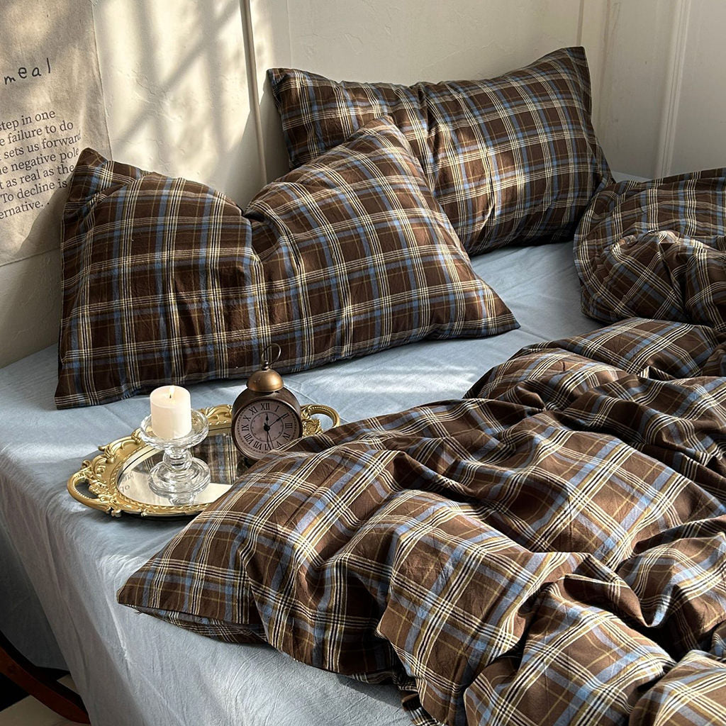 Brown plaid bedding set with matching pillows and duvet on a bed, styled with a candle, alarm clock, and gold tray in a warm, sunlit room.